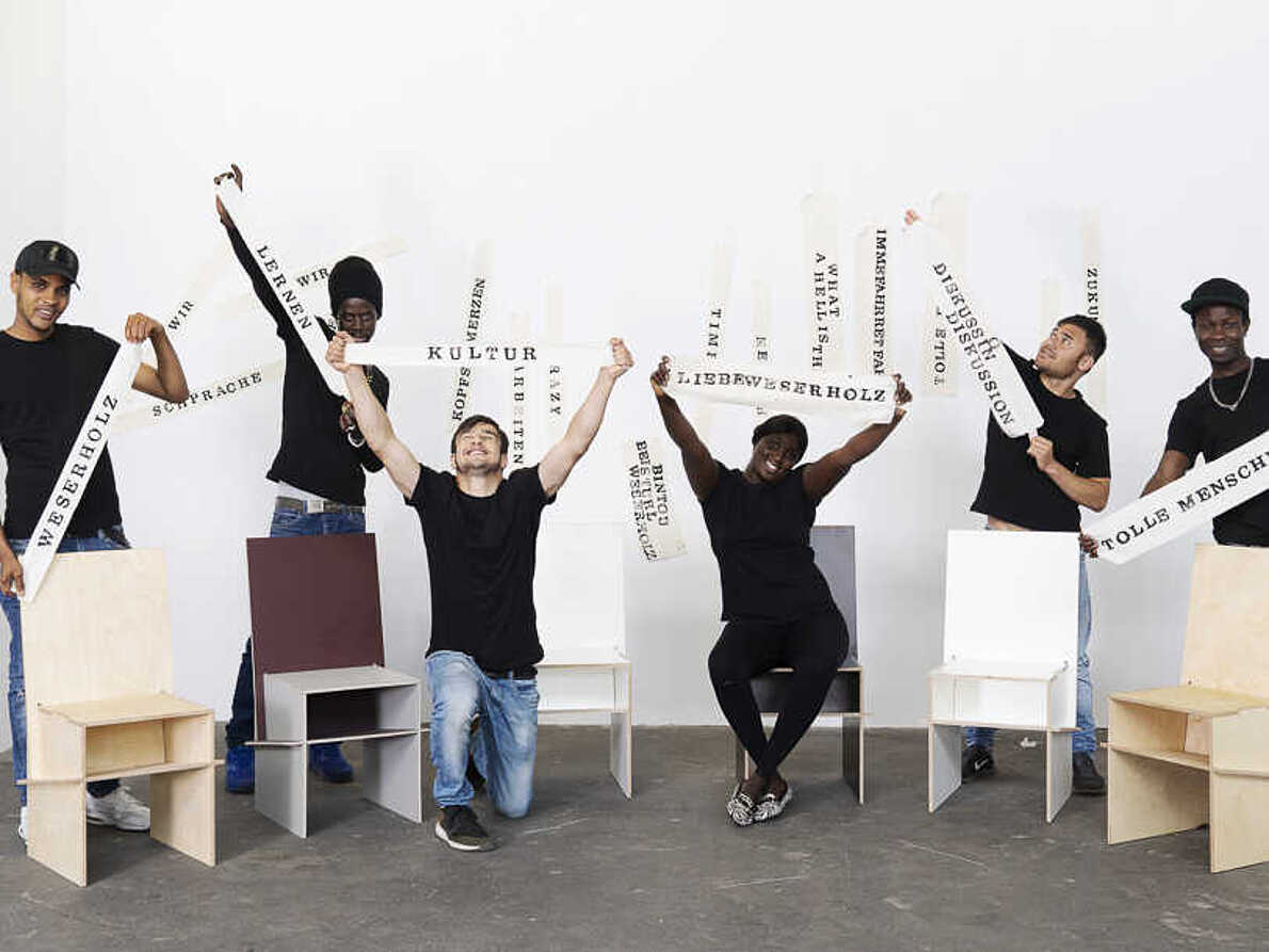 Sechs Personen halten Banner mit Aufschriften wie "Tolle Menschen" "Kultur" und "Lernen" hoch. Vor und hinter ihnen stehen besondere Stühle, der bintou Stuhl von weserholz