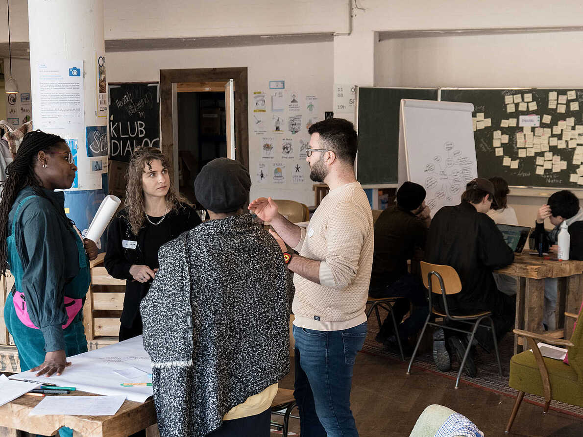 Workshopsituation: Vier Personen stehen um einen Tisch mit Arbeitsmaterialien und unterhalten sich.
